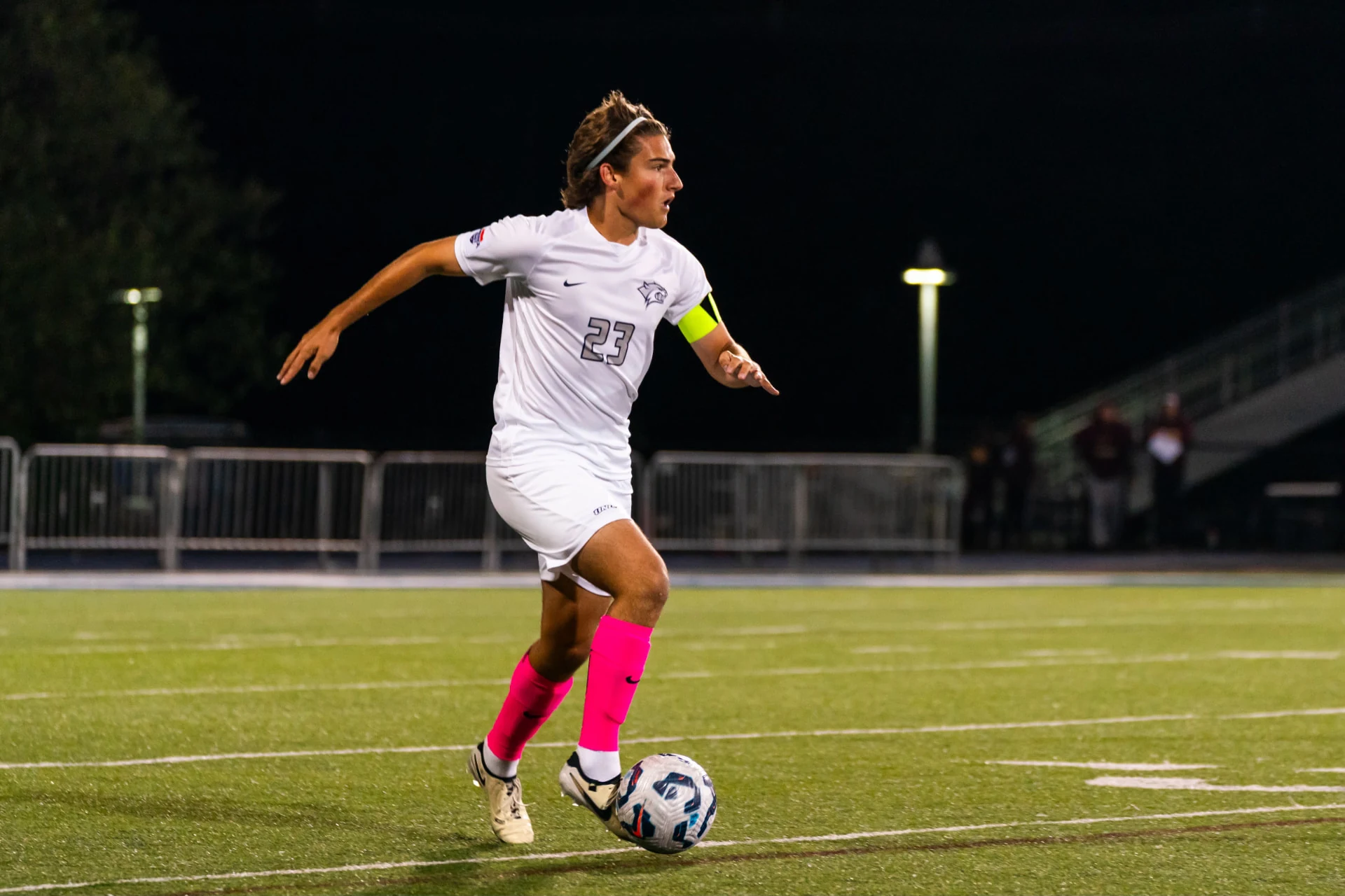 Adison “Adi” Hicks, in his white number 23 University of New Hampshire jersey, striking the ball during a nighttime match.